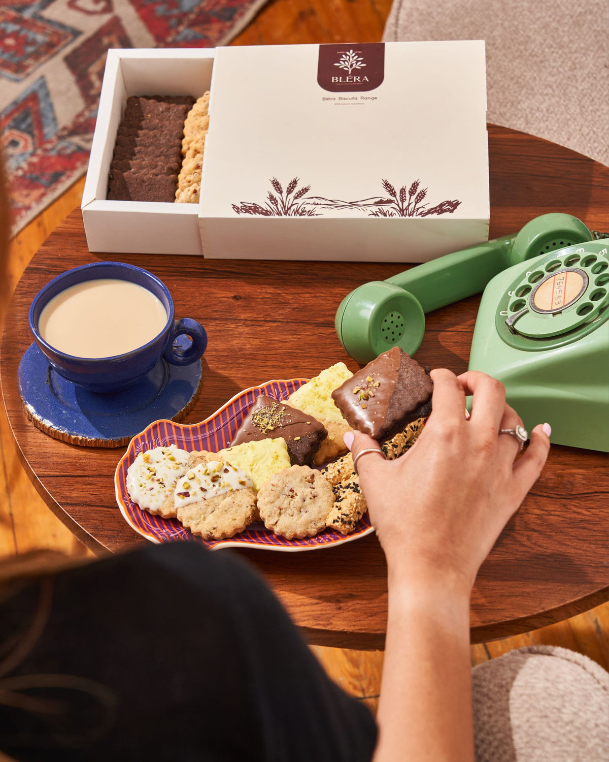 An open Bléra box beside a cup of tea and a plate of biscuits, a hand reaching for one